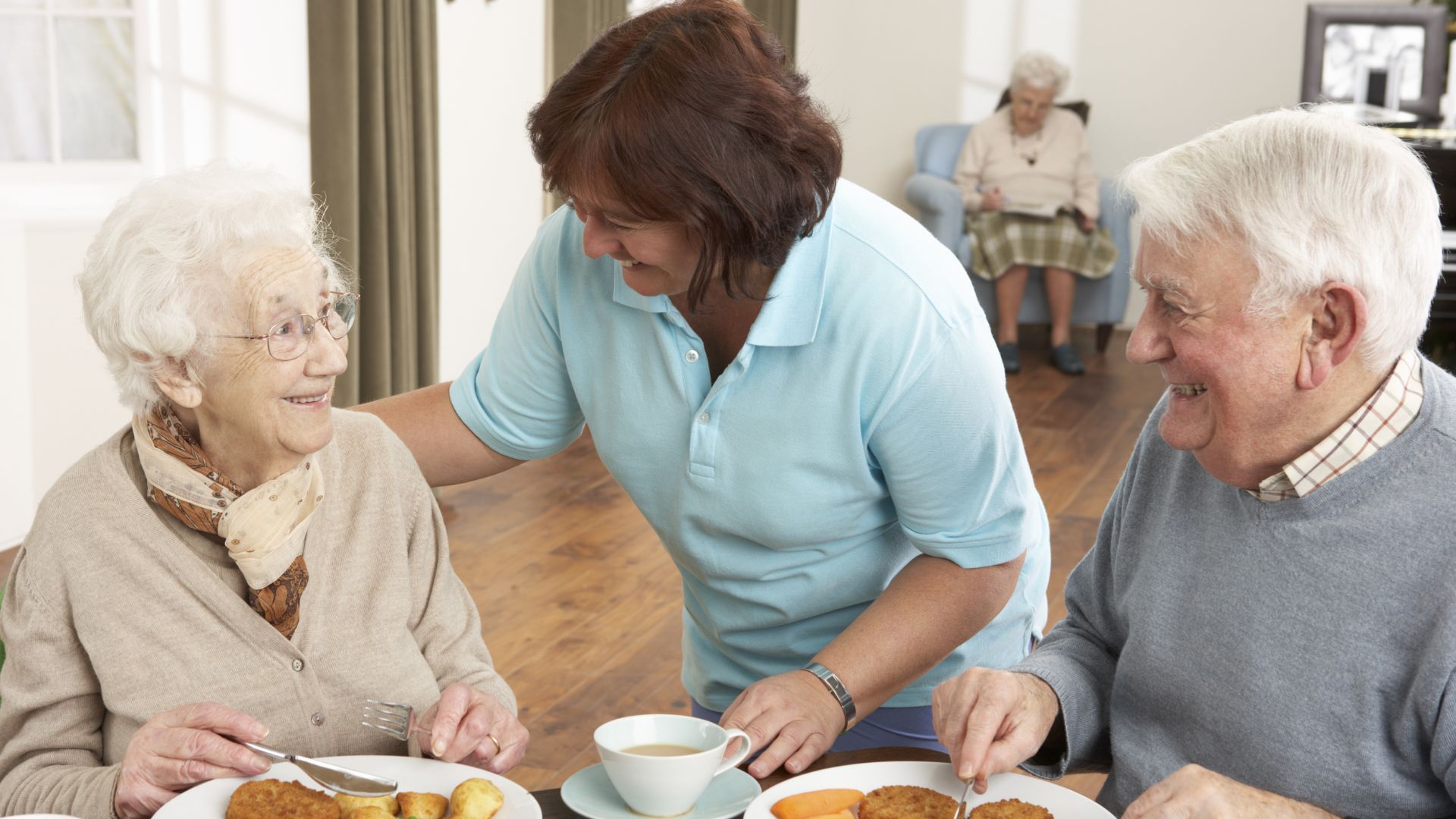 Caregiver serving meals to smiling seniors in a warm, family-style Residential Care home.