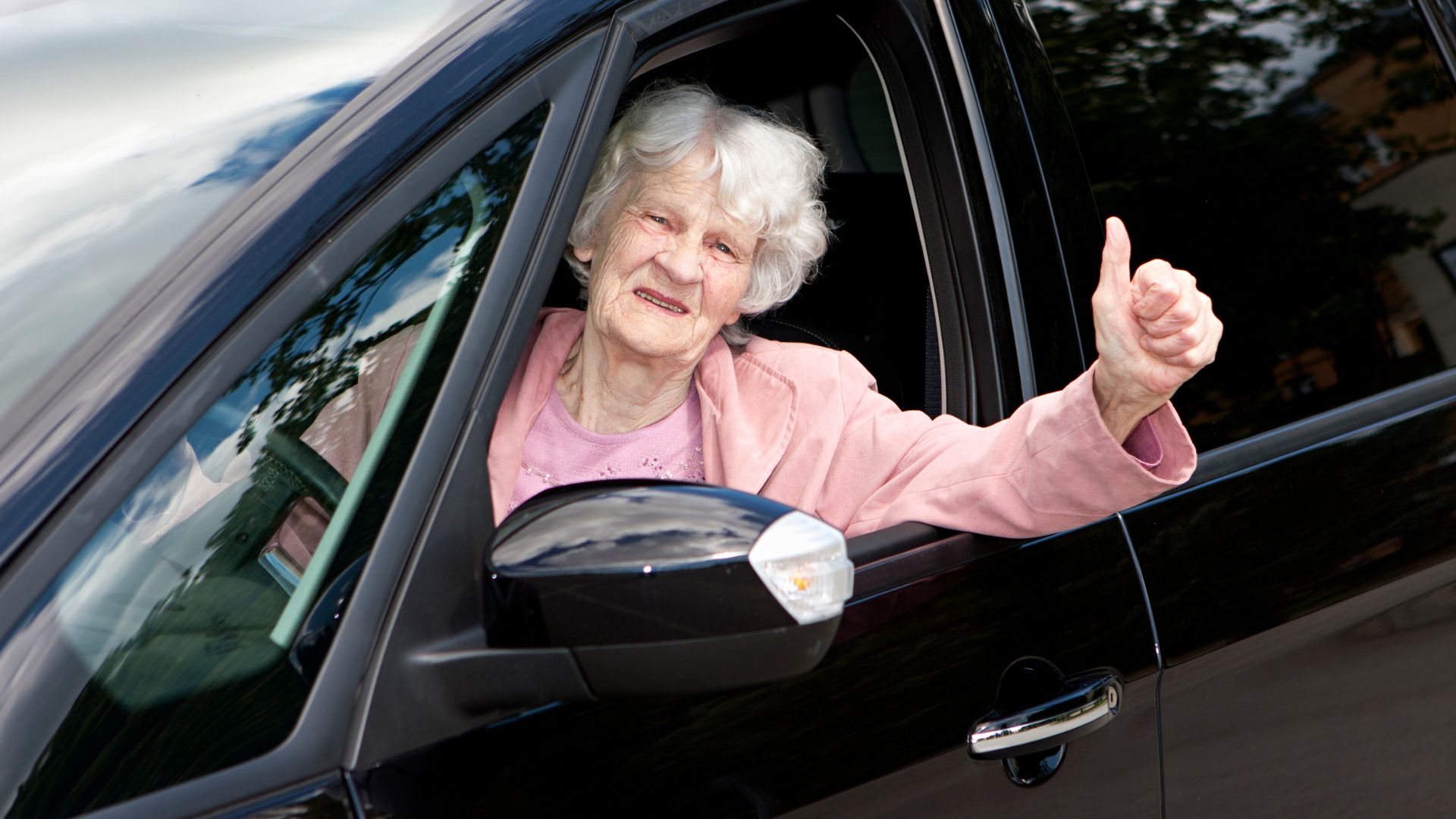 Senior woman giving a thumbs up from her car, representing independence and the importance of programs that go beyond driving with dignity.